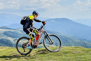 Cyclist man riding electric mountain bike outdoors. Male tourist biking along grassy trail in the mountains, wearing helmet and backpack. Concept of sport, active leisure and nature.
