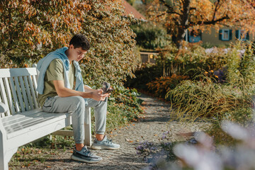 Fototapeta premium A Teenager Sits On A Bench In The Autumn Park Drinks Coffee From A Thermo Mug And Looks Into A Phone. Portrait Of Handsome Cheerful Guy Sitting On Bench Fresh Air Using Device Browsing Media Smm