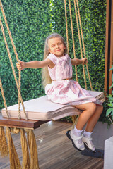 A cute smiling Caucasian girl with curls in a light pink dress swings on a swing. The background is a living green wall. Funny. Happy childhood concept