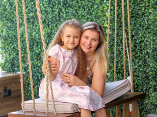 Loving Mom and daughter close-up, Caucasians with blond hair sitting on a hanging swing. The background is a living green wall. Happiness motherhood concept