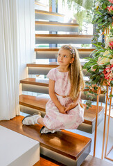 Cute Caucasian girl with blond hair and pink pastel dress sitting on the stairs with floral decoration. Spring decor in the house. Cozy home concept