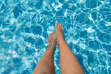 Women's feet dip into the blue water in the pool. Swimming pool. Vacation resorts in summer holidays