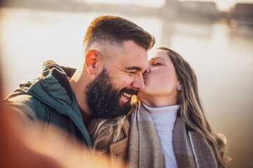 Close up image of happy couple taking selfie outdoor. Woman kissing her  man in cheek.