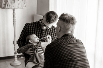 Male gay couple with adopted baby girl at home - Two handsome dads feed the baby girl on kitchen -...