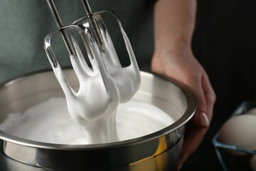 Woman making whipped cream with hand mixer on black background, closeup