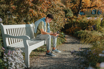 Fototapeta premium A Teenager Sits On A Bench In The Autumn Park Drinks Coffee From A Thermo Mug And Looks Into A Phone. Portrait Of Handsome Cheerful Guy Sitting On Bench Fresh Air Using Device Browsing Media Smm