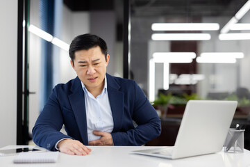 Man has severe stomach pain, nausea and food poisoning, businessman inside office at workplace, worker in business suit working with laptop.