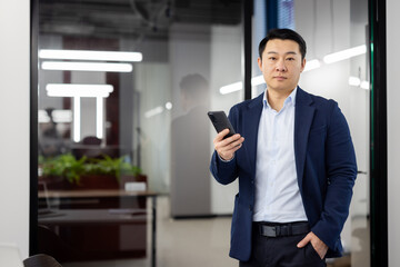 Portrait of serious concentrated businessman asian, man with phone in hands looking at camera, mature boss inside office standing near window, using app on smartphone.