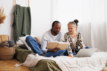 Young diverse couple reading book together near laptop on bed at home