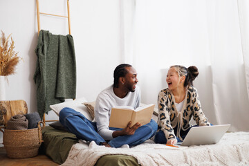 Laughing young multiethnic couple looking at each other while using laptop and reading book on bed...