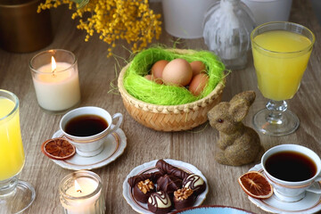 Easter eggs in the basket, bowl of cookies, chocolate pralines, Easter bunny figurine, cups of tea, glasses of juice, flowers and lit candles on the table. Selective focus.