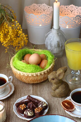 Easter eggs in the basket, bowl of cookies, chocolate pralines, Easter bunny figurine, cups of tea, glasses of juice, flowers and lit candles on the table. Selective focus.