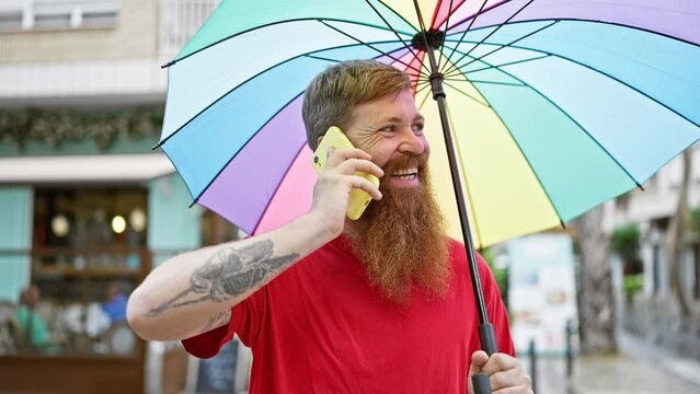 Cheerful young redhead man, confidently enjoying a funny chat on his smartphone, looking up smiling from under his umbrella on a city street.