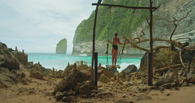 Bikini beach girl model in black swimsuit having fun on swing on on tropical sea shore on summer vacations.