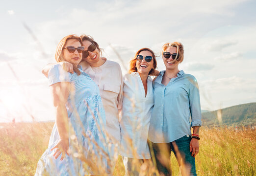 Portrait of four cheerful smiling and laughing women embracing during outdoor walking by high green grass hill. They looking at the camera. Woman friendship, relations, and happiness concept image.