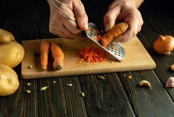 Fotobehang Grating carrots with a grater on a kitchen board. Chef hands preparing vegetable dishes in tavern kitchen © Виталий Борковский