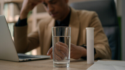 Overworked sick African American business man throw painkiller tablet in glass of water apply fizzy pill with bubbles in drink on office table suffer headache hangover ill businessman prepare remedy 