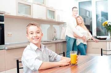 Smiling caucasian boy with a glass of juice looking at camera in the kitchen while his parents cooking dinner at the background.