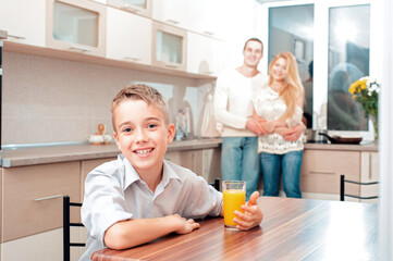 Obraz na płótnie Canvas Smiling boy with a glass of juice looking at camera in the kitchen and his parents standing at the background.
