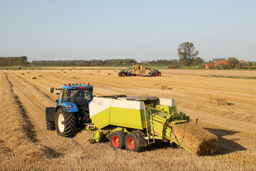 a rural landscape with a tractor and a big baler in front at a straw field and tractors with front loaders in the background