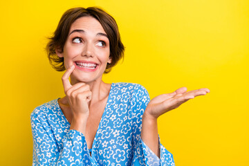 Portrait of dreamy nice girl wear blue blouse look empty space finger on teeth arm hold product isolated on yellow color background