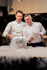 two chefs in white uniforms are preparing liquid nitrogen in the kitchen which spreads all over table
