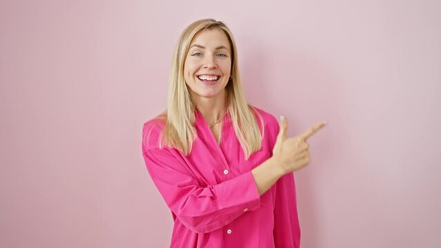 Cheerful young blonde woman smiling and pointing upwards on a pink isolated background