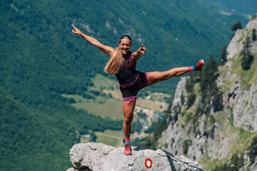 A fit skyrunner is standing on a rock on mountain cliff and balancing on one foot.