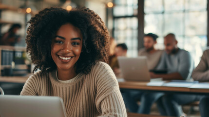 A smiling woman is sitting at a desk with a laptop in a well-lit coworking space, surrounded by other professionals also working on their devices.