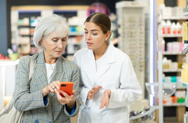 Fototapeta na wymiar Concerned old female customer asking young chemist about product reflected on her phone in large pharmacy