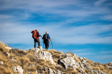 Two hikers walk on the mountain path, helping themselves up the climb with trekking poles.