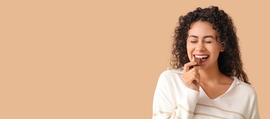 Beautiful young African-American woman eating sweet chocolate on beige background with space for text
