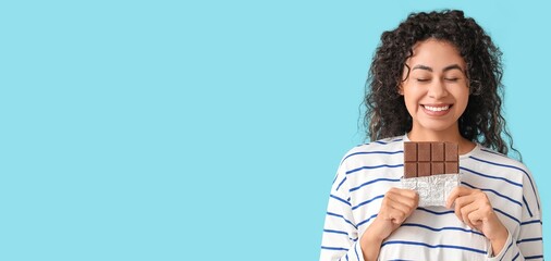 Happy young African-American woman holding sweet chocolate bar on light blue background with space...