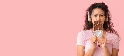 Worried young African-American woman with headphones and sweet chocolate bar on pink background...
