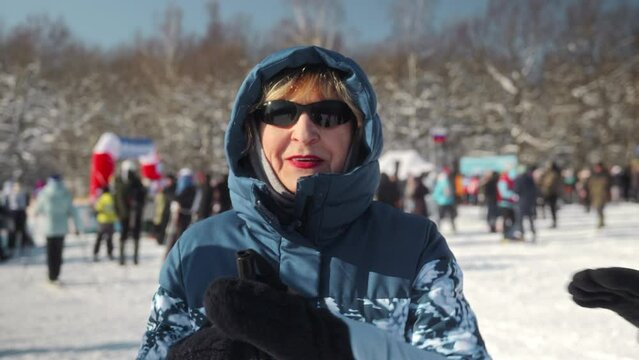 Portrait of an elderly female skier who stands in front of the camera on a sunny day with glasses and ski poles. An elderly woman at a city skiing competition. High quality 4k footage