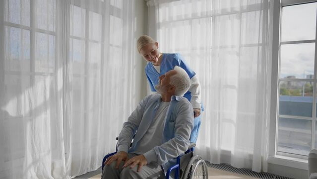 Social worker helps man in wheelchair. Female social welfare physical therapist looks after senior male, talking, providing medical care at home. Living with legs paralysis, elderly trauma concept.