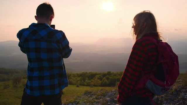 Adolescent boy taking photos of a fantastic mountain landscape at sunset with a smartphone while standing in the viewpoint in the company of a girl with a backpack, handheld shot.