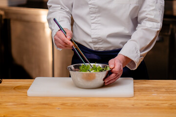 close-up of a chef in a white uniform in a professional kitchen stirring salad with large tweezers
