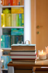 Stack of books, reading glasses and lit candles on the table. Rainbow bookshelf in the background. Selective focus.