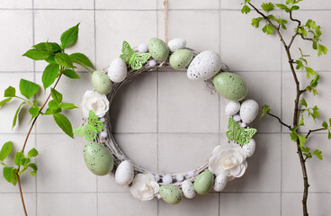 Easter wreath made of flowers, eggs and butterflies with branches hanging on white wall