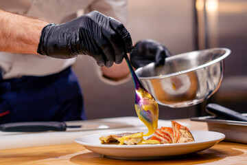 close up chef in a white tunic holding a bowl and pouring sauce from spoon onto the finished dish - obrazy, fototapety, plakaty