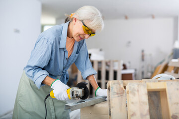 Focused mature woman in protective glasses cutting metal profile using grinding machine, renovating...