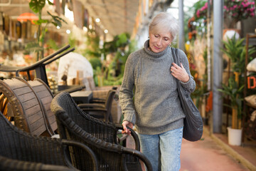 Positive aged female shopper choosing resin wicker chairs for using in outdoor patio or garden in...
