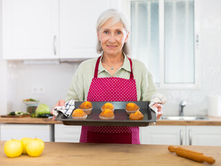 Smiling senior woman in apron holding sheet pan with just baked cupcakes. Old woman baking cakes at...