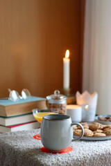 Fototapeta na wymiar Cup of tea or coffee, stack of books, e-reader, glasses, various cookies, almonds, orange juice and lit candles on the table. Hygge at home. Selective focus.
