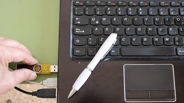 Trabajo remoto en la casa. El hombre que trabaja con la computadora portatil, con el pendrive y tomando notas con el bol&iacute;grafo blanco, forma un videoclip original con un fondo de teclado negro