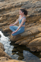 woman sitting on rocks meditating and practicing yoga, reflections in water.