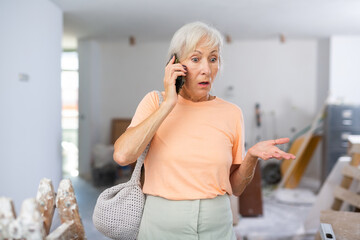 Displeased elderly woman talking on mobile phone in a room being renovated