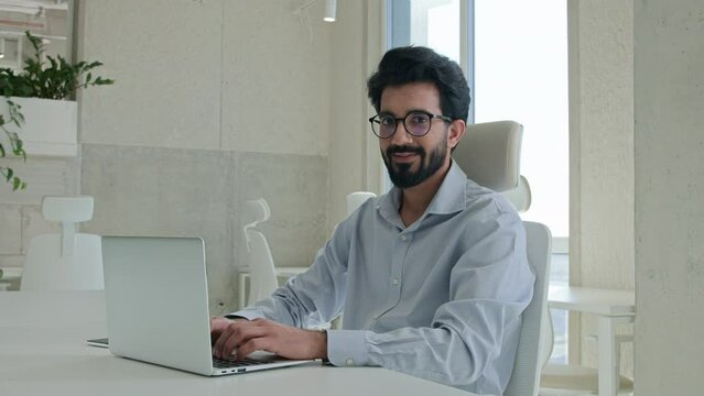 Arabian Indian muslim businessman ethnic man working with computer at corporate office cheerful happy manager employee programmer typing laptop browsing website business work looking at camera smile