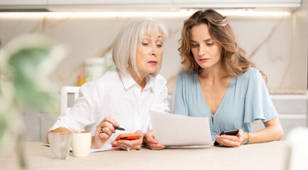 Elderly woman and adult woman discussing agreement while sitting at table in kitchen
